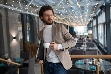 pleased cheerful young man wearing fashion jacket, jeans, roll neck holding cup of coffee and shut laptop in city cafe, looking aside, away, success, free time, spare time