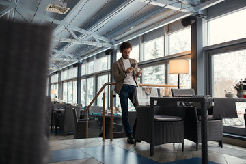 young man in fashion clothes standing at the cafe, drinking tea and dowloading music, full length...