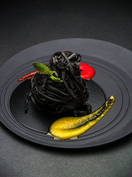 Black spaghetti with cuttlefish ink, basilico sprig, red and yellow souces on round black plate on black background.  Plate service