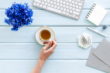 Spring inspiration concept. Work desk with cornflowers bouquet - hand with coffee - on blue wooden background top-down