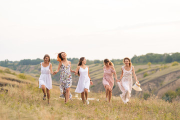 The company of cheerful female friends have a great time together on a picnic in a picturesque place overlooking the green hills. Girls in white dresses dancing in the field