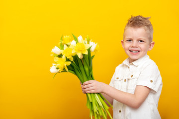 Cheerful little blonde boy gives bouquet of white tulip and daffodil flowers on yellow studio background. gifts flower delivery, Spring Woman day.