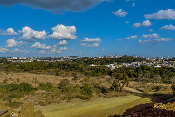 Estrada de terra com vista para cidade no horizonte. Zona urbana envolta por bosques verdes.	