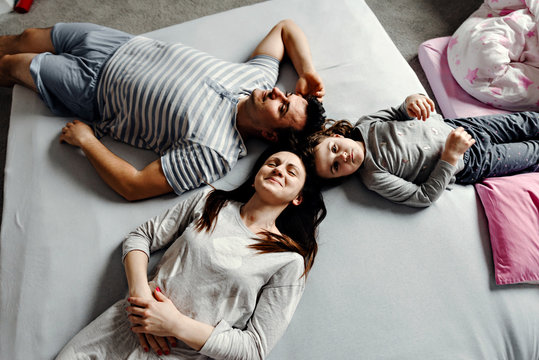 Top view of a cute little girl and her beautiful young parents looking at the camera and smiling, lying on the floor at home,Vvew from above of resting family lying on the floor and looking at camera