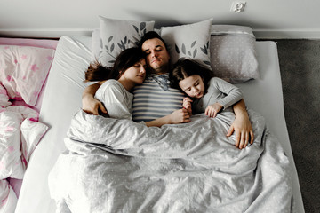 daughter with parents in bed,dad gently kisses his wife and daughter,strong family hugs,family weekend in bed,sleeping family,daughter sleeps with young parents in bed