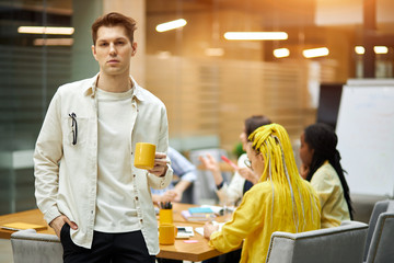 serious young awesome man holding a mug and posing to the camera, free time, spare time, his colleagues sitting at the table and discussing problem