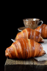 Golden croissants of yeast dough and a cup of coffee on the edge of the wooden table against black background