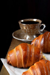 Golden croissants of yeast dough and a cup of coffee on the edge of the wooden table against black background