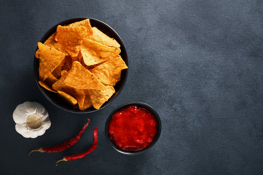 Mexican nachos in a black bowl with hot salsa dip sauce, garlic and chili peppers on textured black background
