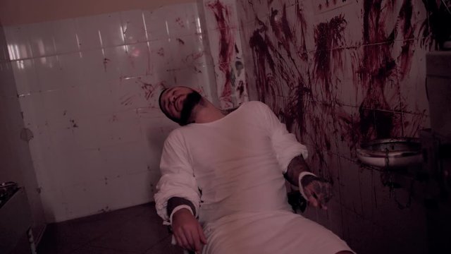 Crazy man in straitjacket tied to a chair, killer psychopath in a room with bloodied walls overdose, death