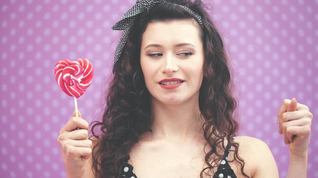 Lovely young curly girl standing, holding sweet red lollipop on the stick in one hand, showing zilch by another hand.