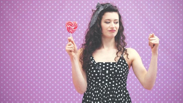 Lovely young curly girl standing, holding sweet red lollipop on the stick in one hand, showing zilch by another hand.
