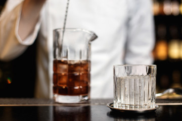 A horizontal image of a rocks glass with ice and bartender stirring a cocktail in a mixing glass with a bar spoon
