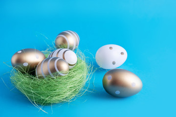 Golden eggs and white Easter eggs painted with gold paint lie in a basket of green grass and are scattered nearby. Easter holiday spring. Blue background. Place for text. Postcard
