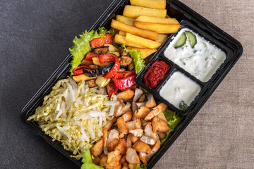Traditional doner kebab in a black plastic container, sour cream, fried potato, roast meat, sauces, salad, cabbage and roasted vegetables on grey background