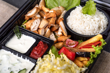 Traditional doner kebab in a black plastic container, sour cream, white boiled rice, roast meat, chiken, sauces, salad, cabbage and roasted vegetables on grey background