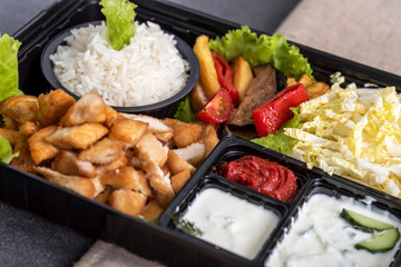 Traditional doner kebab in a black plastic container, sour cream, white boiled rice, roast meat, chiken, sauces, salad, cabbage and roasted vegetables on grey background