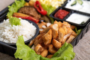 Traditional doner kebab in a black plastic container, sour cream, white boiled rice, roast meat, chiken, sauces, salad, cabbage and roasted vegetables on grey background