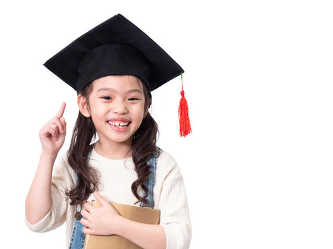 Asian Little Cute Girl 6 Years Old Wearing Graduation Hat And Holding A Book On Hands. Preschool Lovely Kid With A Book And Pointing Finger Up Isolated On White Background With Clipping Path.