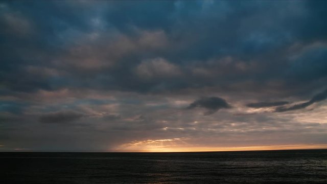 A dramatic time-lapse of a sunset over open ocean water, sun beams leak through the clouds in the distance illuminating the sky as the sun sets the scene gets darker at the end the sun reveals itself