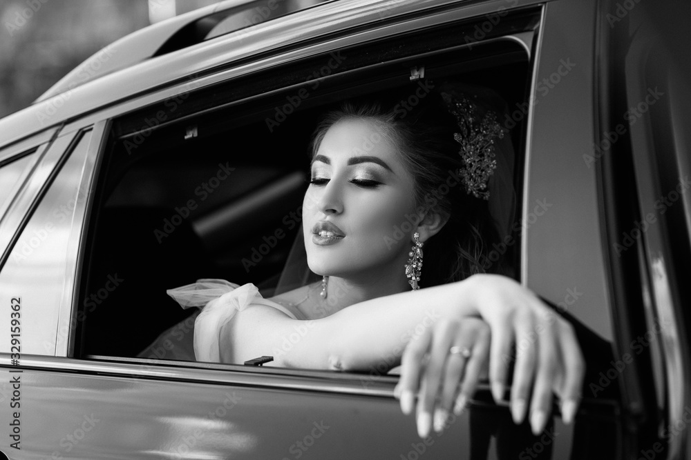 Wall mural Gorgeous elegant bride posing in stylish black car, sitting inside in saloon. Luxury wedding in vintage style. Portrait. Black white photo