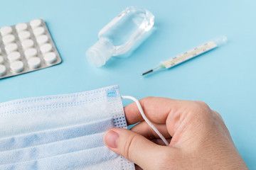 Woman hand holds disposable surgical mask over a blue surface with thermometer, pills and...