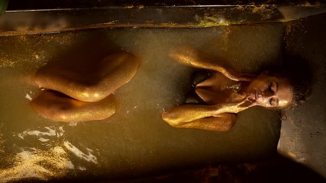  excited woman in bath caresses her breasts, top view, girl with golden skin lies in water stroking her body