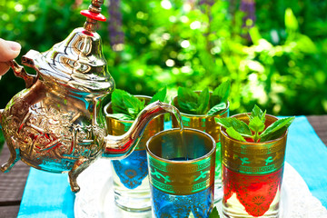 Pouring of moroccan mint tea from a cooper teapot into a glass cup in a sunny garden on a wooden table.