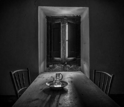 Comedor De Casa Abandonada  Con Mesa Y Sillas A Contraluz De Una Ventana Medio Abierta.