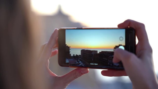 Beautiful colorful sunset at the sea beach through the smartphone screen, back view of girl holding modern cellphone and taking pictures of summer sundown. Abstract lights and colors, technology and