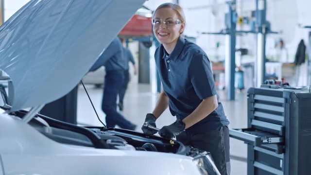 Beautiful Empowering Female Car Mechanic is Working on a Vehicle in a Service. She Looks Happy While Using a Ratchet. Specialist is Wearing Safety Glasses. She Looks at a Camera and Smiles.