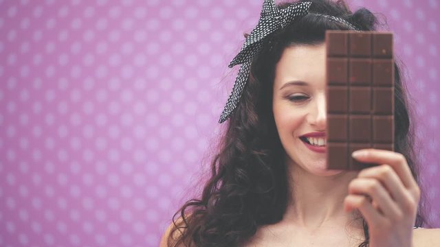 Pretty young lady with curly black hair and headband in polka-dots, hiding her eye behind a bar of chocolate.