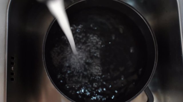 Black stainless cooking pan lying in the sink and pure water flowing in it. Water votex and bubbles on the surface, stream of water from the kitchen tap, creating droplets from high water pressure