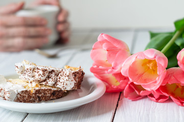 Slice of biscuit tiramisu cake, pink tulips and female hands with cup of coffee