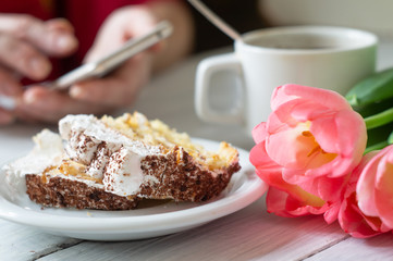 Tiramisu cake, female hands holding smartphone and cup of coffee and pink tulips