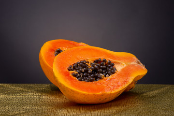 Bright vibrant orange papaya fruit sliced in half on a gold coloured surface with black seeds piling out