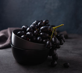 a cluster of juicy black grapes lies in a dark gray bowl on a dark gray table