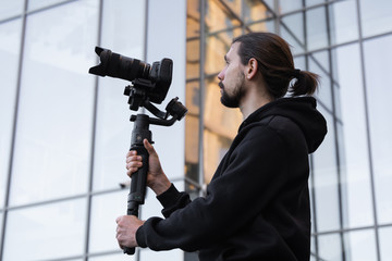 Young Professional videographer holding professional camera on 3-axis gimbal stabilizer. Pro equipment helps to make high quality video without shaking. Cameraman wearing white hoodie making a videos.