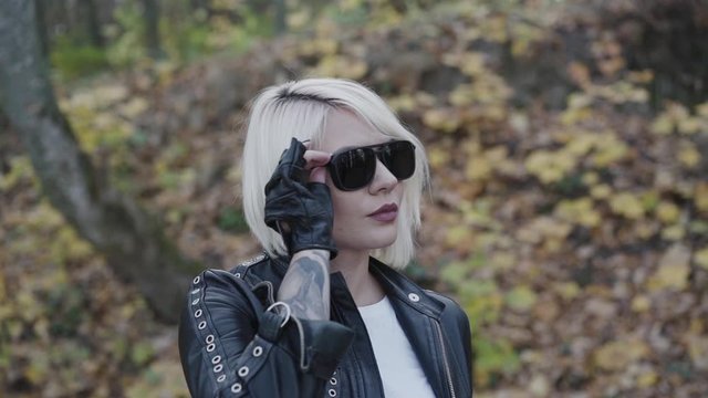 Portrait of glamorous female biker corrects sunglasses and looks in park