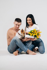 Obraz na płótnie Canvas Close up portrait young couple sit and kissing, holding yellow flowers and dog in studio on white background. couple embracing with dreamy amorous expression. Lovely family. Celebrating woman's day.