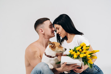 Close up portrait young couple sit and kissing, holding yellow flowers and dog in studio on white background. couple embracing with dreamy amorous expression. Lovely family. Celebrating woman's day.