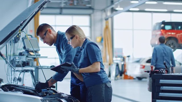 Two Mechanics in a Service are Inspecting a Car After They Got the Diagnostics Results. Female Specialist is Comparing the Data on a Tablet Computer. Repairman is Using a Ratchet to Repair the Faults.