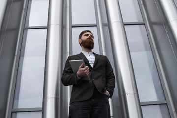 Modern Young bearded Business man standing with a digital tablet. Young hipster businessman holding tablet in hands outdoor. Working online with a tablet while standing outside on an office building.