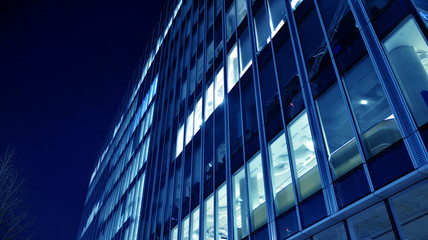 Naklejka na ściany i meble Night architecture - building with glass facade.Blue color of night lights. Modern building in business district. Concept of economics, financial. Photo of commercial office building exterior. Abstra