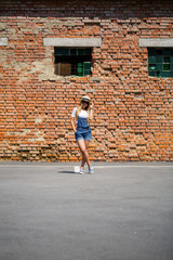 Girl in denim overalls, a white T-shirt and a light hat on a background of a brick building under the bright sun