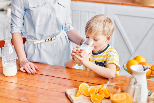 The Boy Drinks Milk From A Glass. Mother And Son Are Smiling While Having A Breakfast In Kitchen. Mom Is Pouring Milk Into Glass