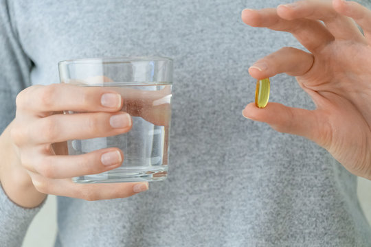 woman in a gray jacket is holding an omega-3 fish oil capsule and a glass of water. Biologically active additive. Vitamin E, a Golden tablet against beriberi.