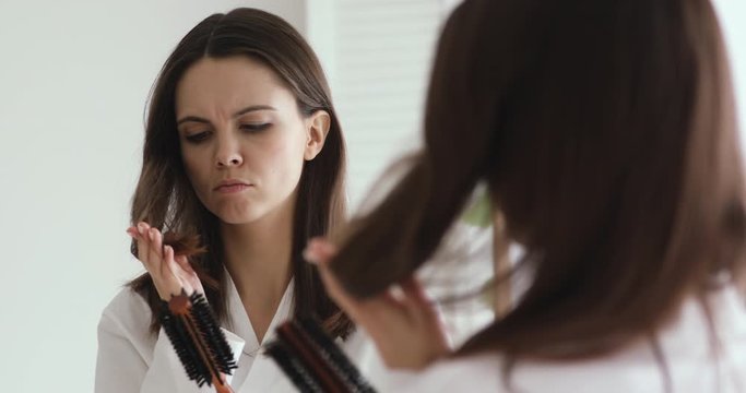Depressed young woman brushing looking at split ends reflecting in bathroom mirror. Worried girl feels upset about brittle damaged dry hair loss concept. Women hormone problems or vitamins deficiency