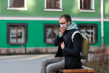 Man in glasses feeling sick, sitting on bench, coughing, wearing protective mask against transmissible infectious diseases and protection against virus - covid-19. New coronavirus 2019-nCoV from China