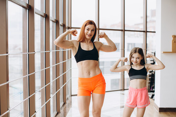 Plakat Beautiful strong woman and charming little daughter are showing their biceps and smiling while working out at gym